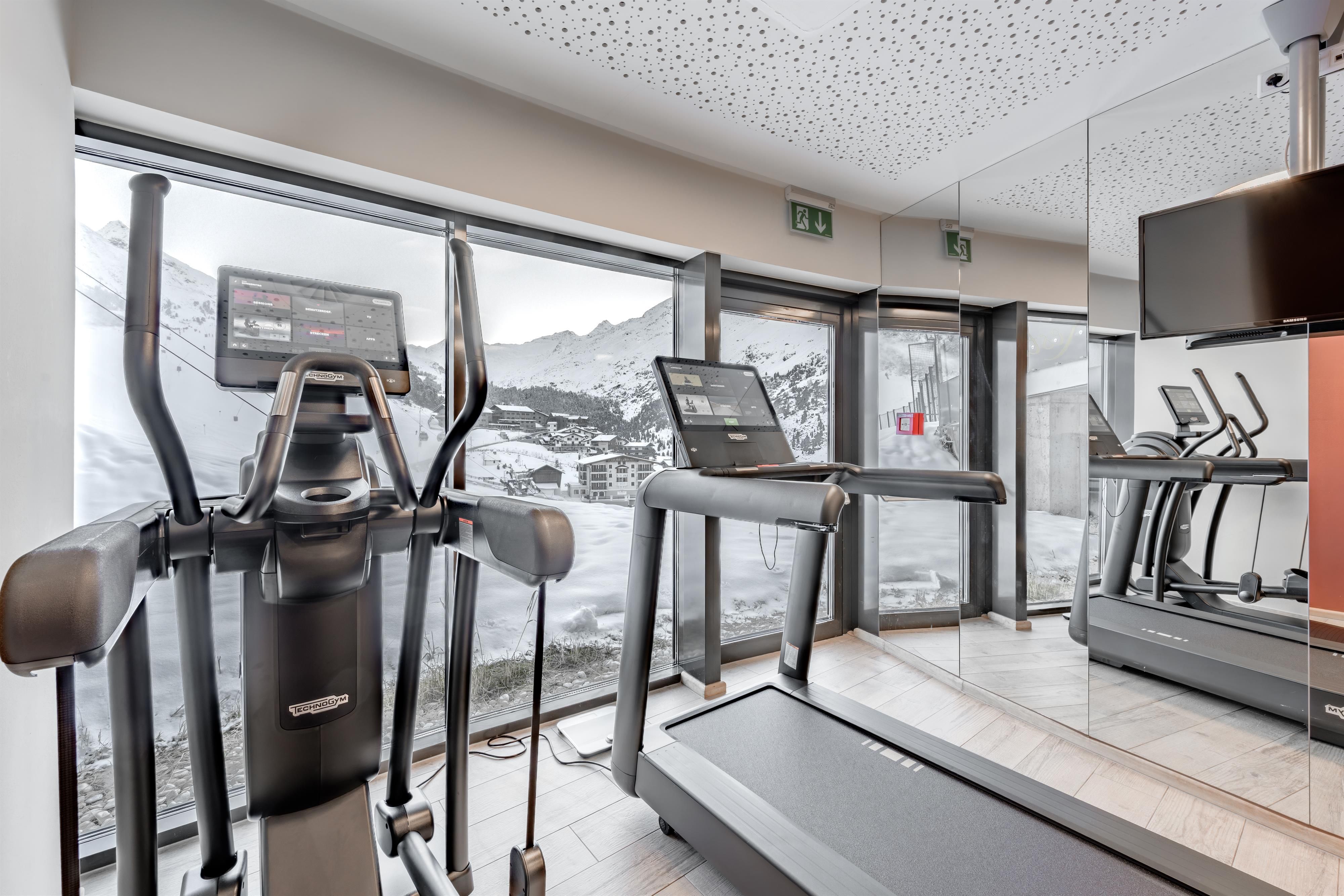 A modern fitness room with a view of the mountains. In front of a large window stand a treadmill and an elliptical trainer.