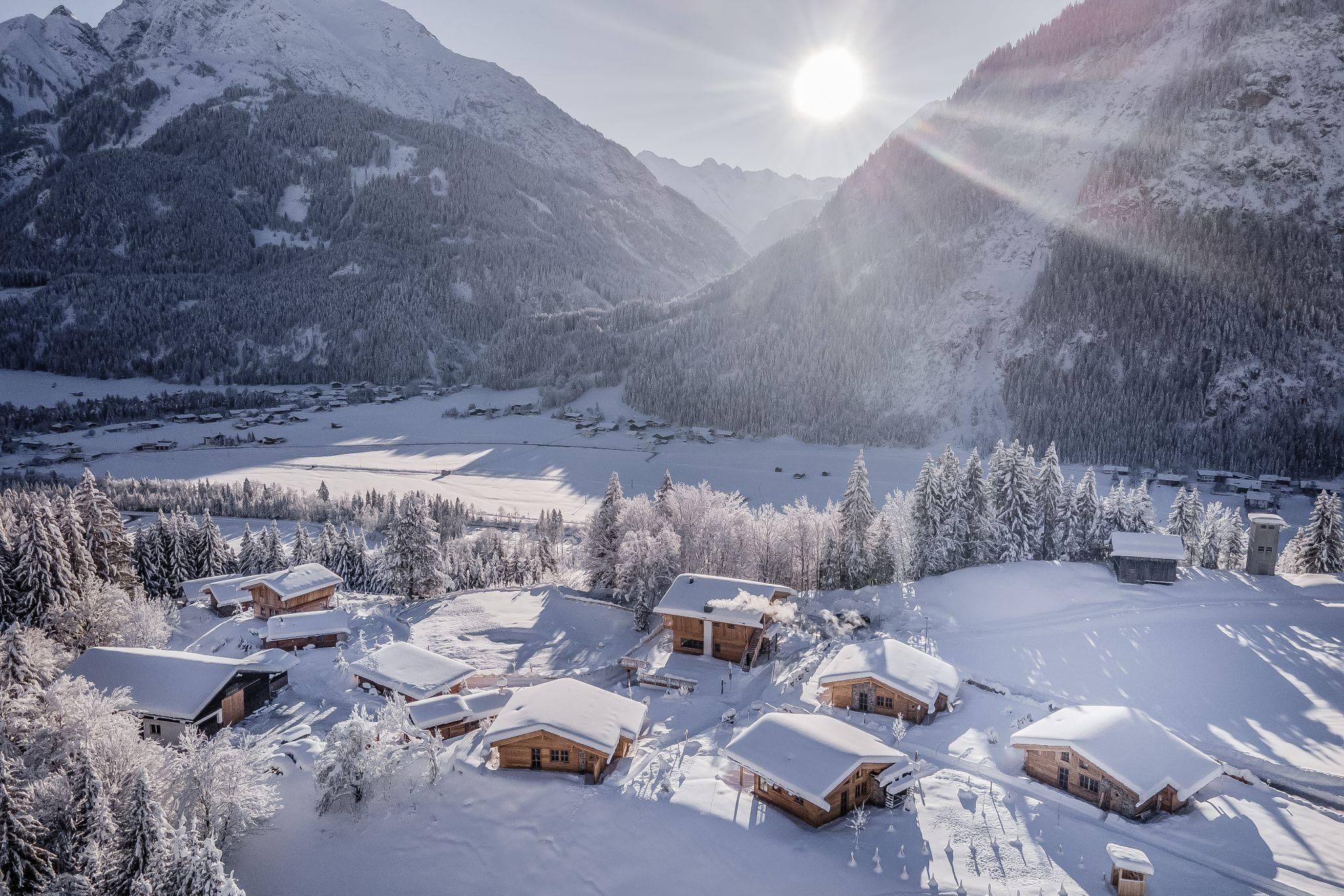 A picturesque winter landscape with snow-covered chalets and tall mountains. The sun shines brightly over the serene valley.