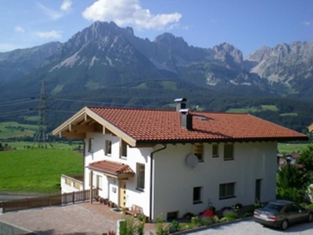 A beautiful house with a red roof in a green landscape. In the background, majestic mountains rise under a blue sky.
